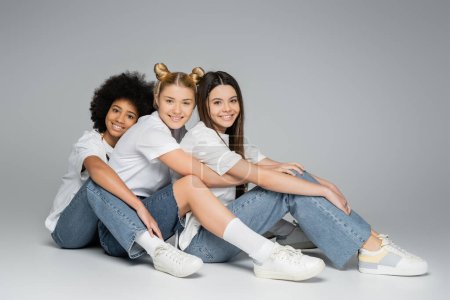 Téléchargez les photos : Pleine longueur de copines adolescentes souriantes et multiethniques en t-shirts blancs et jeans bleus regardant la caméra tout en étant assis et posant sur fond gris, concept de modèles adolescentes multiethniques - en image libre de droit