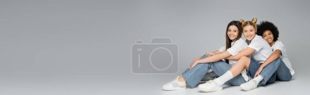 Full length of positive teen multiethnic girlfriends in casual t-shirts and jeans looking at camera while sitting on grey background, multiethnic teen models concept, banner with copy space