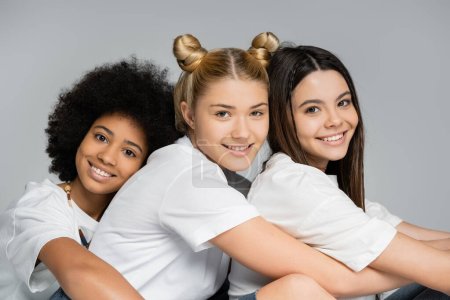 Portrait of smiling and interracial girls in casual white t-shirts looking at camera together and sitting next to each other isolated on grey, multiethnic teen models concept, friendship and bonding