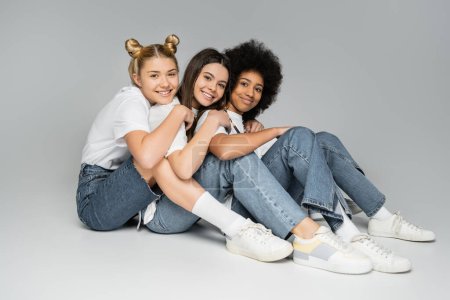Full length of teen and cheerful multiethnic girlfriends in white t-shirts and jeans hugging and posing together on grey background, multiethnic teen models concept, friendship and bonding