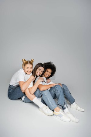 Volle Länge positiver und jugendlicher multiethnischer Freunde in lässigen weißen T-Shirts, Jeans und Turnschuhen, die in die Kamera schauen und auf grauem Hintergrund sitzen, Konzept multiethnischer Teenager-Modelle