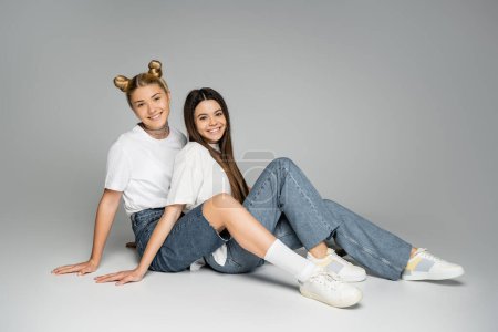 Smiling blonde and brunette teenage girls in white t-shirts, jeans and sneakers looking at camera while sitting together on grey background, teen models concept, friendship and bonding