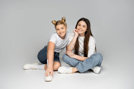 Positive brunette and blonde teenage girls in white t-shirts, jeans, denim shorts and sneakers looking at camera while sitting on grey background, teen models concept, friendship and bonding