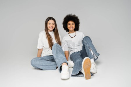 Full length of positive african american teenage girl in white t-shirt and blue jeans sitting next to brunette girlfriend on grey background, lively teenage girls concept, friendship and bonding