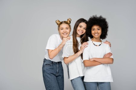 Photo for Positive and teenage girls in white t-shirts and jeans hugging confident african american girlfriend crossing arms while standing isolated on grey, lively teenage girls concept, friendship and bonding - Royalty Free Image
