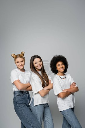 Positive and multiethnic teen girlfriends in stylish white t-shirts and jeans looking at camera and crossing arms while standing together isolated on grey, lively teenage girls concept