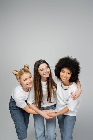 Cheerful and multiethnic teenage girlfriends in white t-shirts and jeans hugging brunette friend together and standing isolated on grey, lively teenage girls concept, friendship and bonding