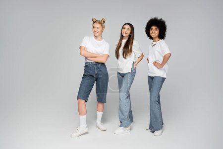 Full length of smiling interracial teen girls in white t-shirts and jeans posing and looking at camera while standing on grey background, lively teenage girls concept, friendship and companionship