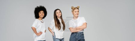 Joyful and multiethnic teenage girlfriends in white t-shirts posing and looking at camera while standing next to each other isolated on grey, lively teenage girls concept, banner 