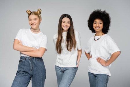 Adolescentes positives et multiethniques en jeans et t-shirts blancs décontractés posant et souriant à la caméra tout en étant isolées sur le concept gris, adolescent vif, amitié et camaraderie
