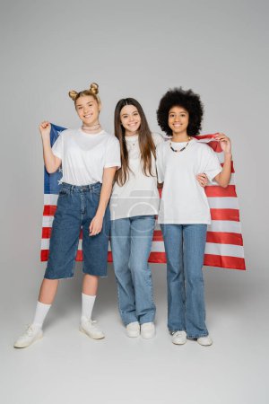 Volle Länge multiethnische Teenie-Freundinnen in weißen T-Shirts und Jeans, die eine ameikanische Fahne in der Hand halten und in die Kamera auf grauem Hintergrund schauen, lebhafte Teenager-Mädchen, Freundschaft und Kameradschaft.