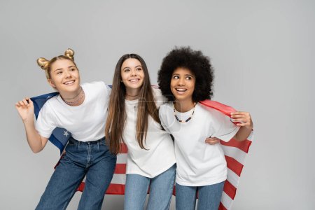 Teenage and multiethnic girlfriends in white t-shirts and jeans smiling away while holding american flag and standing on grey background, energetic teenage friends spending time, friendship