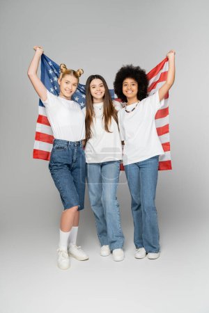 Volle Länge positiver und multiethnischer Teenager-Mädchen in weißen T-Shirts mit amerikanischer Flagge und Blick in die Kamera auf grauem Hintergrund, energische Teenager-Freunde, die Zeit verbringen, Freundschaft