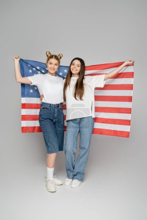 Full length of cheerful blonde and brunette teen girlfriends in white t-shirts holding american flag and standing on grey background, energetic teenage friends spending time, friendship 