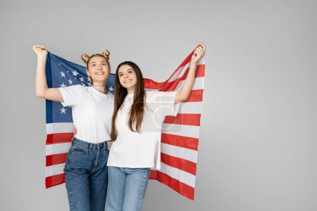 Brünette und blonde Teenager-Freundinnen in weißen T-Shirts und Jeans, die die amerikanische Flagge in der Hand halten und aufblicken, während sie isoliert auf grauen, energischen Teenagerfreunden stehen, die Zeit verbringen, Freundschaft