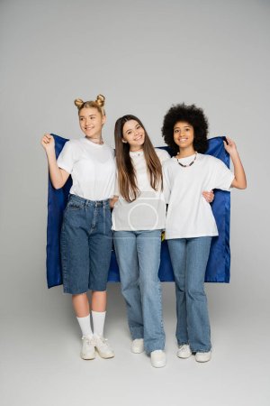 Pleine longueur de copines adolescentes souriantes et multiethniques en jeans et t-shirts blancs tenant le drapeau bleu et debout sur fond gris, des amis adolescents énergiques passent du temps