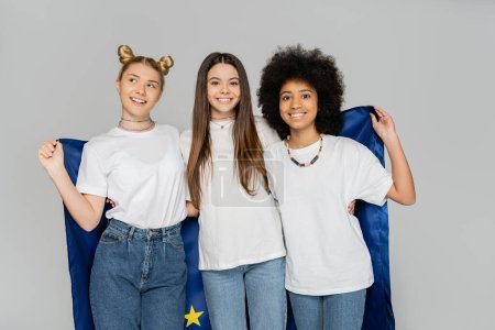 Cheerful and multiethnic teen girlfriends in white t-shirts and jeans holding european flag together and standing isolated on grey, energetic teenage friends spending time, friendship 