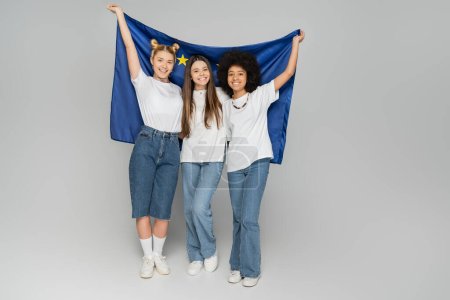 Full length of cheerful interracial teen girlfriends in white t-shirts holding European flag together and standing on grey background, energetic teenage friends spending time