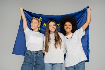 Positive und multiethnische Teenager-Mädchen in weißen T-Shirts und Jeans mit blauer Europaflagge, während sie isoliert auf grauen, energiegeladenen Teenagerfreunden stehen und Zeit miteinander verbringen