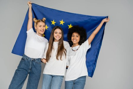 Excited and smiling multiethnic teenagers in white t-shirts and jeans holding blue european flag while posing and standing isolated on grey, energetic teenage friends spending time