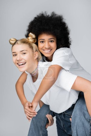 Foto de Alegre afroamericana adolescente chica en blanco camiseta y jeans piggybacking en rubia amigo y divertirse aislado en gris, energético adolescente modelos pasar tiempo, amistad y compañerismo - Imagen libre de derechos