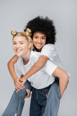 Teen african american girl in jeans and white t-shirt hugging blonde girlfriend while looking at camera and standing isolated on grey, energetic teenage models spending time