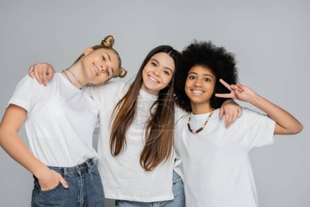 Smiling brunette teen girl in white t-shirt and jeans hugging multiethnic girlfriends posing and gesturing isolated on grey, energetic teenage models spending time, friendship and companionship