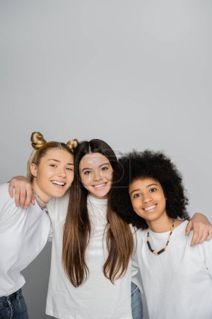 Smiling brunette teen girl in white t-shirt hugging interracial girlfriends and looking at camera together isolated on grey, energetic teenage models spending time, friendship and companionship