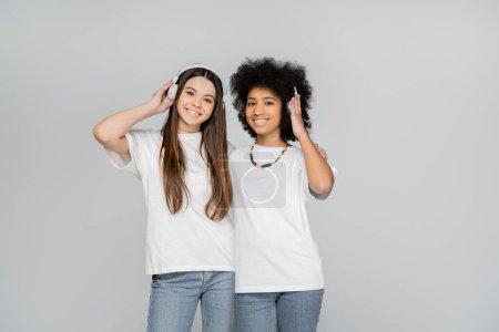 Joyful and interracial teen girls in white t-shirts and jeans listening music in headphones and looking at camera isolated on grey, energetic teenage models spending time, friendship and companionship