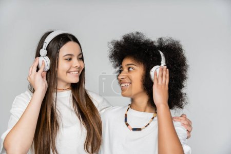 Fröhliches brünettes Teenie-Mädchen in weißem T-Shirt umarmt afrikanisch-amerikanische Freundin mit Kopfhörern isoliert auf grau, energische Teenager-Models verbringen Zeit, Freundschaft und Kameradschaft
