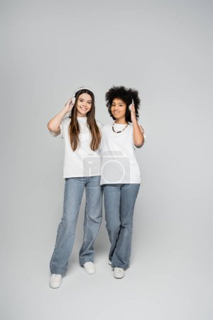 Foto de Longitud completa de novias multiétnicas y adolescentes en camisetas blancas y jeans escuchando música en auriculares y de pie sobre fondo gris, modelos adolescentes enérgicos pasar tiempo - Imagen libre de derechos