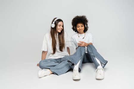 Smiling and multiethnic teen girlfriends in white t-shirts and jeans using headphones and smartphone while sitting on grey background, teenagers bonding over common interest