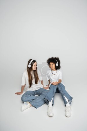 Cheerful multiethnic teenage girlfriends in jeans and white t-shirts listening music in headphones and using smartphone on grey background, teenagers bonding over common interest mug #662018166