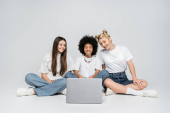 Joyful and multiethnic teen friends in white t-shirts and jeans looking at laptop together and sitting on grey background, teenagers bonding over common interest, friendship and companionship tote bag #662018260