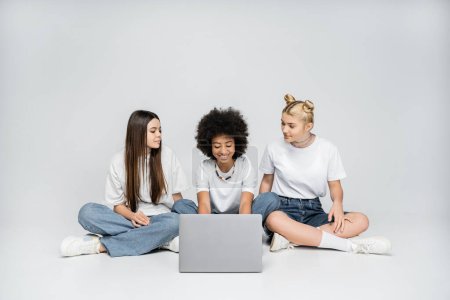 Lächelndes afrikanisch-amerikanisches Mädchen mit Laptop in der Nähe von Freundinnen in weißen T-Shirts und Jeans, die zusammen auf grauem Hintergrund sitzen, Teenager, die sich über gemeinsame Interessen zusammentun