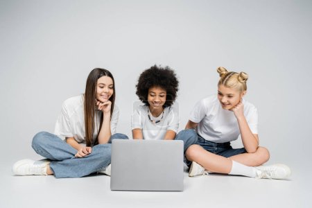 Téléchargez les photos : Adolescente afro-américaine utilisant un ordinateur portable près de copines en t-shirts et jeans blancs tout en étant assis ensemble sur fond gris, les adolescents collant sur l'intérêt commun, l'amitié et la camaraderie - en image libre de droit