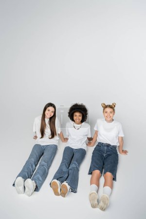 Vue grand angle de copines adolescentes souriantes et multiethniques en t-shirt blanc et jeans regardant la caméra tout en étant assis sur fond gris, modèles d'adolescence et concept de génération z