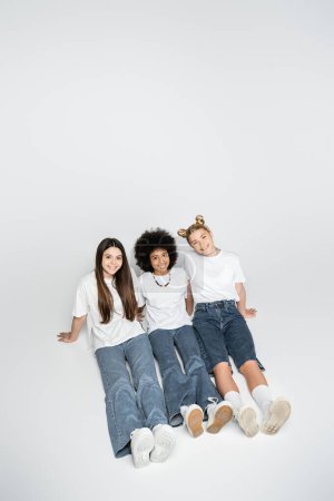 High angle view of positive and multiethnic teenage friends in white t-shirts and jeans looking at camera and sitting together on grey background, adolescence models and generation z concept