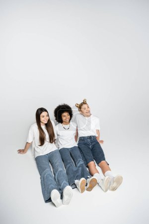 High angle view of positive interracial teenage girlfriends in white t-shirts and blue jeans looking away while sitting on grey background, adolescence models and generation z concept