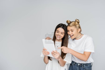 Smiling blonde teen girl in jeans and white t-shirt pointing with finger at digital tablet and hugging brunette friend isolated on grey, teenagers bonding over common interest, Touch Screen