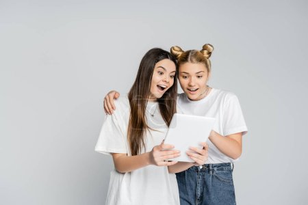 Excited teenage girlfriends in white t-shirts using digital tablet together while standing isolated on grey, teenagers bonding over common interest, friendship and companionship