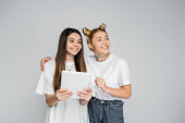 Smiling blonde teenage girl in white t-shirt and jeans hugging brunette girlfriend with digital tablet and looking away together isolated on grey, teenagers bonding over common interest Tank Top #662018388