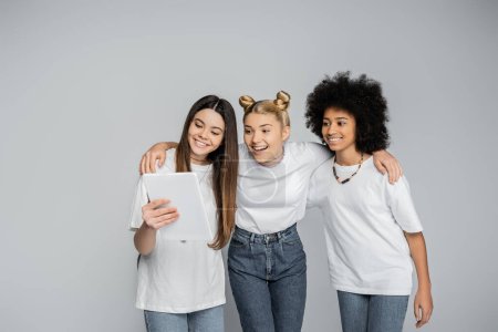 Positive blonde teenager in white t-shirt and jeans hugging multiethnic girlfriends and using digital tablet while standing isolated on grey, teenagers bonding over common interest