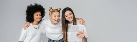 Smiling interracial teenage girlfriends in white t-shirts using digital tablet together while hugging and standing isolated on grey, teenagers bonding over common interest, banner 