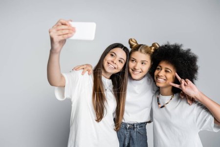 Foto de Sonrientes novias adolescentes con camisetas blancas abrazando y haciendo gestos mientras se toman selfie en un teléfono inteligente sobre un fondo gris, adolescentes que se unen por el interés común, la amistad y la compañía - Imagen libre de derechos