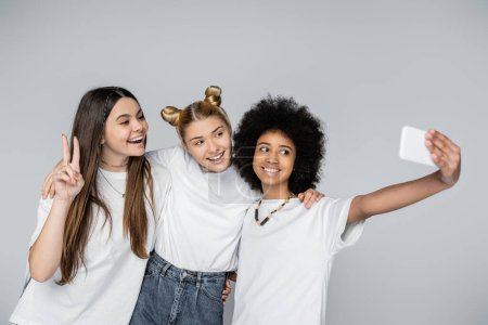 Lächelnde afrikanisch-amerikanische Teenagerin in weißem T-Shirt, die ein Selfie auf dem Smartphone macht, mit Freundinnen, die sich isoliert auf grau umarmen, Teenager, die sich über gemeinsame Interessen, Freundschaft verbinden 