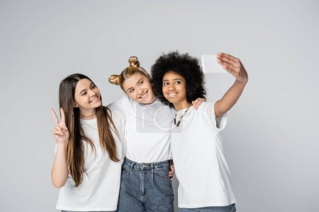 Lächelnde multiethnische Teenager-Freunde in weißen T-Shirts, die ein Selfie mit dem Smartphone machen, sich umarmen und gestikulieren, während sie isoliert auf grau stehen, Teenager, die sich über gemeinsame Interessen, Freundschaft bekennen 