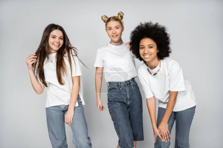 Cheerful and interracial teenage girlfriends in blue jeans and white t-shirts posing together and looking at camera isolated on grey, adolescence models and generation z concept