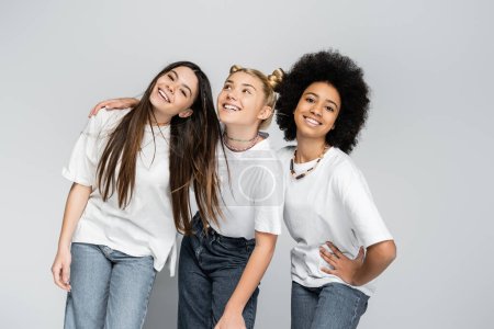 Foto de Novias adolescentes multiétnicas en jeans y camisetas blancas casuales posando mientras se abrazan y se paran juntas aisladas en modelos grises, adolescentes y concepto de generación z, amistad - Imagen libre de derechos