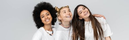 Positive und multiethnische Teenager-Freundinnen in lässigen weißen T-Shirts, die sich umarmen und in die Kamera schauen, während sie isoliert auf grauen, pubertären Modellen und dem Konzept der Generation Z stehen, Banner 